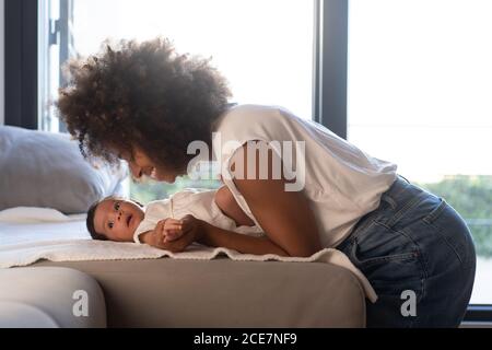 Vue latérale d'une femme afro-américaine enchantée jouant avec un bébé mignon sur le canapé à la maison Banque D'Images