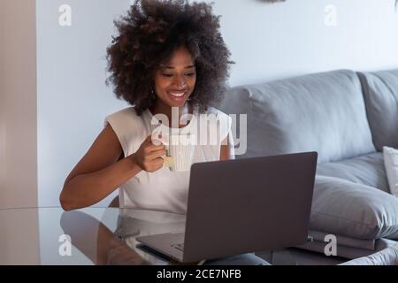 Femme entrepreneure afro-américaine concentrée assise à table avec tasse de thé et de travail à distance tout en utilisant netbook à la maison Banque D'Images