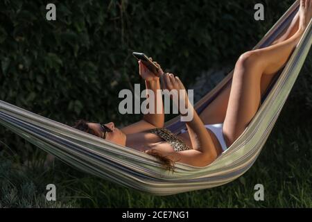 Vue latérale d'une jeune femme gaie en bikini et lunettes de soleil couché dans un hamac près de plantes vertes à l'aide d'un smartphone tout en se reposant en été dans le jardin Banque D'Images