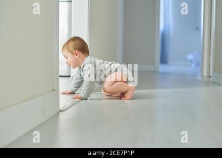 Vue latérale de l'adorable petit bébé en body gris rampant sur le sol dans une chambre confortable à la maison Banque D'Images