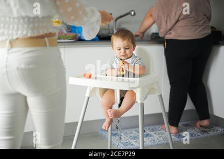 Petit enfant concentré assis sur une chaise haute pour bébé et en train d'examiner avocat frais pendant que la mère prépare la nourriture dans la cuisine maison Banque D'Images