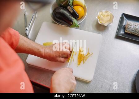 Du dessus de la récolte anonyme chef coupant le zeste de citron frais pendant la cuisson, près de la plaque avec du poisson et des légumes crus sur une surface métallique Banque D'Images