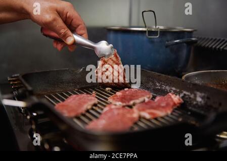 Faites frire des tranches de bœuf fines non reconnaissables sur une poêle à frire sur la cuisinière tout en montrant une délicieuse surface de viande à l'aide de pinces dans la cuisine Banque D'Images