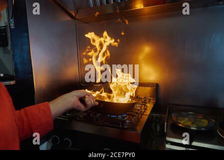 Un chef non reconnaissable est coupé en uniforme tenant une poêle tout en flamant ingrédients dans une poêle à feu lent au restaurant cuisine le soir et vue sur le feu lumineux Banque D'Images