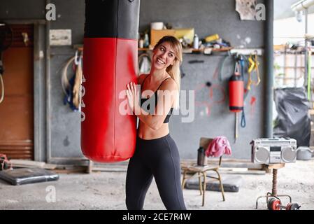 Positive jeune Sportswoman portant des leggings et soutien-gorge de sport enserrant sac de poinçonnage et regardant loin heureux après l'entraînement intense dans le complexe rural garage gym Banque D'Images