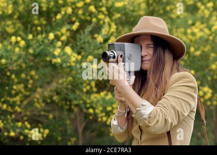 Une photographe féminine au chapeau avec des yeux fermés filmant de la vidéo avec caméra vidéo rétro en se tenant près des arbres avec des fleurs de jaune fleurs à la lumière du jour Banque D'Images