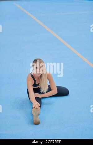Corps complet de jeune femme ciblée dans des vêtements de sport assis sur sol bleu et se pencher vers l'avant tout en étirant le corps pendant entraînement physique sur le terrain de sport Banque D'Images