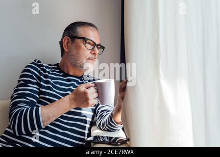 Vue latérale de l'homme barbu qui regarde par les fenêtres tout en dégustant une boisson chaude le matin à la maison Banque D'Images