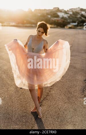 Le corps entier enchante la jeune femme artiste dans une élégante robe de danse avec les yeux fermés dansant dans le dos éclairé dans le carré vide avec ville floue en arrière-plan dans le soleil été coucher de soleil Banque D'Images