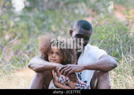 Positif noir homme dans des vêtements décontractés assis avec mignon petit fille sur des tours et s'inclinant sur le tronc d'arbre tout en dépensant journée d'été ensoleillée en forêt Banque D'Images