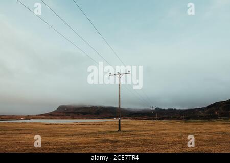 Poteaux électriques situés sur un terrain sec et herbacé en Islande près mer calme entourée de montagnes rocheuses contre le ciel brumeux Banque D'Images
