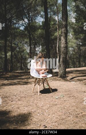 Seule fille pieds nus en robe assis sur un tabouret embrassant les genoux et en regardant vers le bas dans la forêt avec de hauts arbres sous bleu ciel en été Banque D'Images