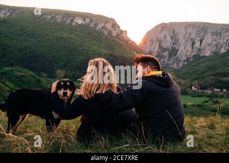 Vue arrière d'un couple amoureux qui s'enserre dans un pré avec une douceur chien tout en se relaxant sur fond de montagnes majestueuses pendant les vacances En Transylvanie Banque D'Images