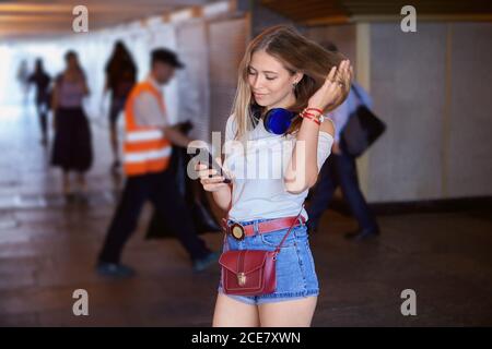 Jeune femme en passe-bas. Belle fille caucasienne environ 20 ans avec de longs cheveux blonds et des écouteurs est debout dans le tunnel avec smartphon Banque D'Images