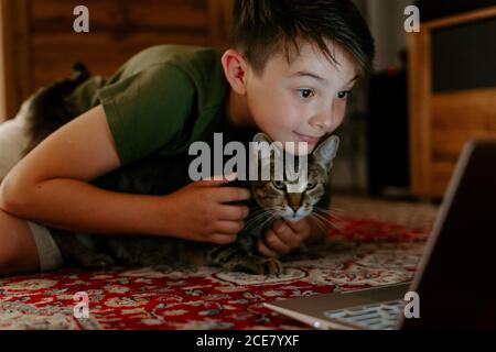 Joyeux adorable enfant qui câline le chat et s'amuser en s'asseyant sur le sol et regarder des vidéos sur un ordinateur portable Banque D'Images