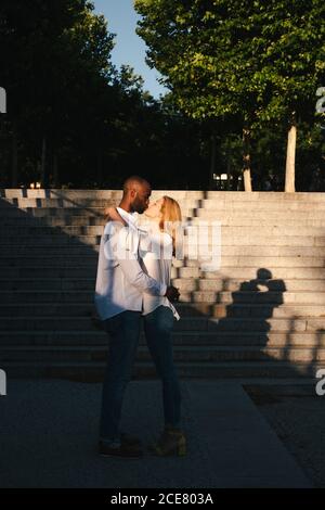 Vue latérale complète du corps avec couple multiethnique embrassant et embrassant en vous tenant face à face contre l'escalier le soir ensoleillé rue à Madrid Espagne Banque D'Images