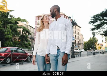 Couple multiracial souriant heureux en jeans et chemises blanches marchant le long du trottoir et en tenant les mains en milieu urbain pendant le soleil jour Banque D'Images