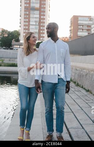 Couple multiracial plein corps heureux souriant en Jean et blanc chemises marchant le long du trottoir et tenant les mains dans un environnement urbain par beau temps Banque D'Images