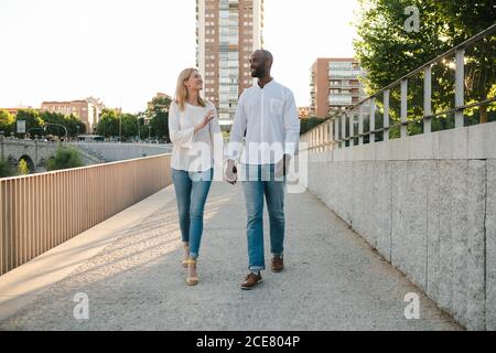 Couple multiracial plein corps heureux souriant en Jean et blanc chemises marchant le long du trottoir et tenant les mains dans un environnement urbain par beau temps Banque D'Images