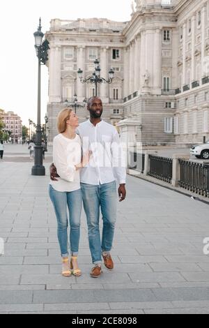 Couple multiracial plein corps gai en jeans et chemises blanches Marcher sur la chaussée le long de la rue à Madrid tout en liant et discussion Banque D'Images