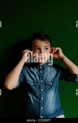 Un écolier portant une chemise en Jean en regardant loin tout en se tenant debout contre la nuit fond vert pour écouter de la musique sur des écouteurs blancs sans fil modernes Banque D'Images