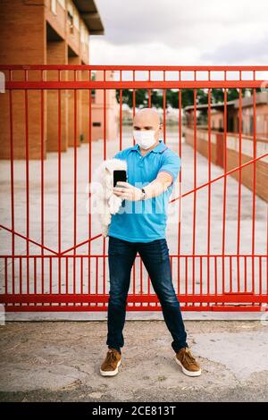 Homme sérieux dans une chemise bleue décontractée et masque debout Avec un joli chien blanc Bichon Frise sur les mains contre le rouge palissade en métal emportant un selfie avec un téléphone portable le jour de l'été Banque D'Images