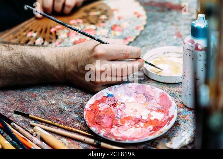 Peintre utilisant un pinceau pour dessiner sur le ventilateur de main en étant assis à une table désordonnée et en travaillant dans un atelier créatif Banque D'Images