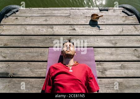 De dessus de jeune homme en chemise rouge couché sur tapis sur le remblai en bois et recréant avec les yeux fermés après séance de yoga Banque D'Images