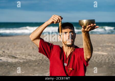 Homme barbu concentré en chemise rouge jouant bol de chant tibétain avant la méditation sur la plage de sable contre ciel bleu sans nuages et océan sans fin Banque D'Images