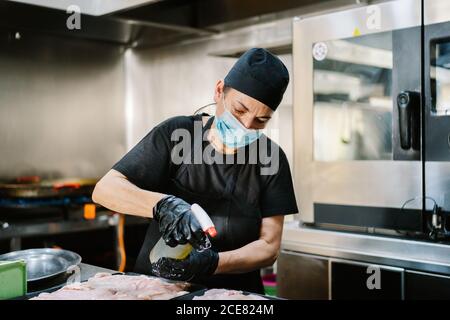 Femme de cuisine concentrée portant des gants en latex et un masque médical pulvérisé avec de la viande de poulet à l'huile placée sur la lèchefrite avant de cuire Banque D'Images