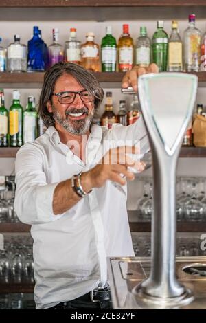 Homme ethnique adulte en chemise blanche versant de l'alcool à partir de la bière colonne dans le verre tout en étant debout dans la barre Banque D'Images