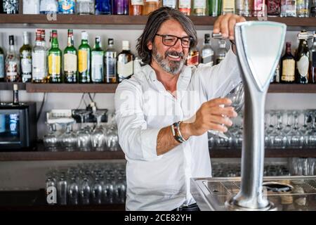 Homme ethnique adulte en chemise blanche versant de l'alcool à partir de la bière colonne dans le verre tout en étant debout dans la barre Banque D'Images