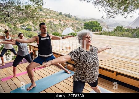 Groupe de plongeurs motivés personnes dans différents âges s'étirant les jambes Et les chevilles pendant l'exécution de Virabhadrasana B pose pendant le yoga en plein air classe Banque D'Images