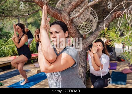 Groupe de femmes actives dans les vêtements de sport équilibrant sur la jambe pendant Pratiquer la position de yoga Garudasana pendant la leçon dans le parc sous le soleil jour Banque D'Images