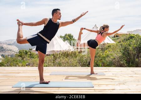 Vue latérale de la pleine longueur flexible et pieds nus homme et femme dans les vêtements de sport pratiquant le yoga tout en faisant le Seigneur de la danse poser debout sur des tapis sur la plate-forme en bois située sur la nature contre les tentes et les montagnes par temps ensoleillé Banque D'Images