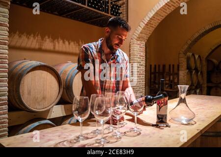 Homme concentré dans une chemise à carreaux décontracté lire des informations sur le vin étiquette de bouteille en vous tenant près de la table avec des verres à vin contre fûts en bois dans un domaine chaleureux Banque D'Images