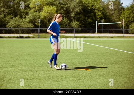 Jeune Sportswoman debout sur un terrain herbeux près du ballon et regardant la caméra au-dessus de l'épaule pendant l'entraînement de football le jour ensoleillé Banque D'Images