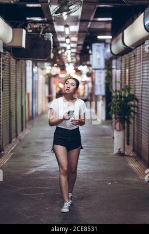 Corps complet jeune femme asiatique dans des vêtements décontractés avec mobile téléphone à la main marchant sur un chemin étroit souterrain piétonnier avec magasins fermés en ville Banque D'Images