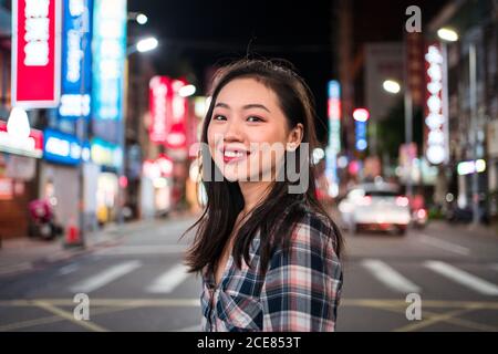 Vue latérale d'une femme asiatique joyeuse du millénaire en chemise à carreaux regarder la caméra avec le sourire en traversant la rue dans moderne quartier illuminé de la ville la nuit Banque D'Images