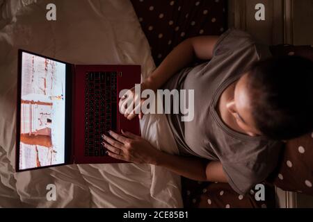 D'en haut, une jeune femme asiatique ethnique méconnue assise sur le lit utilisation du netbook le soir Banque D'Images