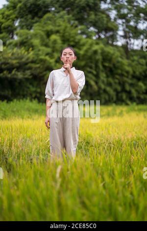 Femme asiatique calme montrant un geste secret avec le doigt sur les lèvres en vous tenant dans le champ de riz et en regardant l'appareil photo Banque D'Images