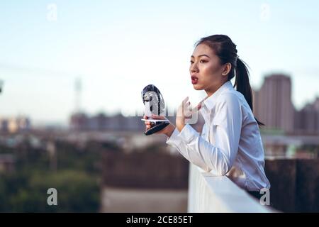 Vue latérale de la jeune femme asiatique dans une tenue habillée à l'aide de mini-ventilateur portable pour se reposer sur le toit en été chaud soirée en ville Banque D'Images