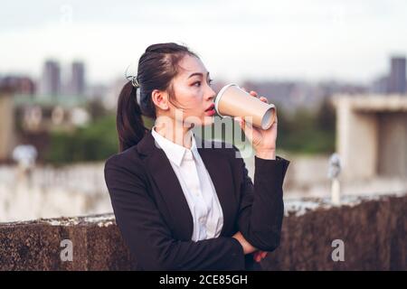 Jeune femme d'affaires asiatique concentrée dans une tenue habillée buvant une tasse de café jetable tout en se tenant sur le toit de l'immeuble de la ville Banque D'Images