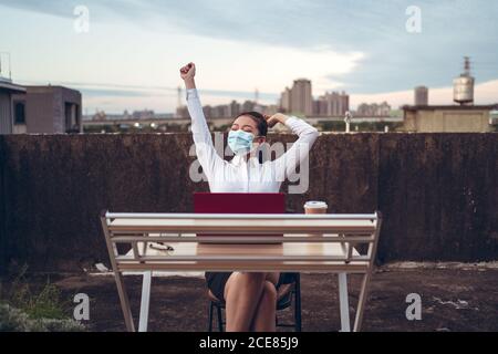 Jeune femme asiatique en tenue formelle et masque de protection table avec ordinateur portable et une tasse de café terrasse sur le toit et détente après un travail à distance pendant une pandémie de coronavirus Banque D'Images