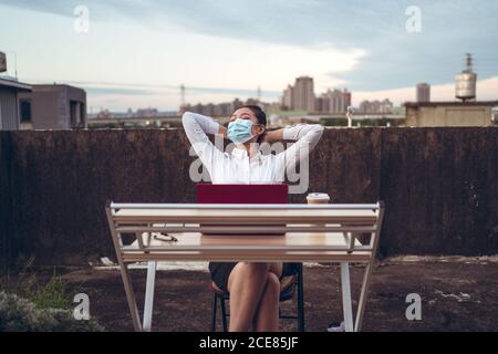 Jeune femme asiatique en tenue formelle et masque de protection table avec ordinateur portable et une tasse de café terrasse sur le toit et détente après un travail à distance pendant une pandémie de coronavirus Banque D'Images