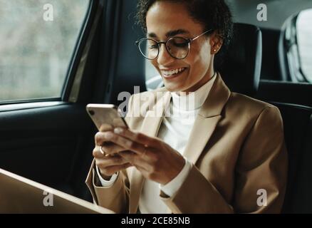 Femme souriante voyageant en voiture. Femme d'affaires assise à l'arrière d'une voiture et lecture du message texte. Banque D'Images