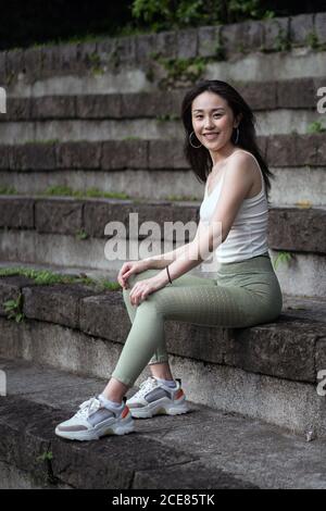 Vue latérale sur toute la longueur sportive asiatique femme porter serré décontracté vêtements reposant sur l'ancien escalier en pierre dans le parc tout en regardant à la caméra heureusement Banque D'Images