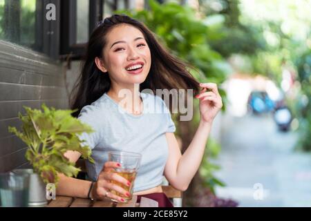 Jeune femme asiatique assise au comptoir avec une boisson fraîche à l'intérieur café et profiter du week-end tout en touchant les cheveux et en regardant appareil photo Banque D'Images
