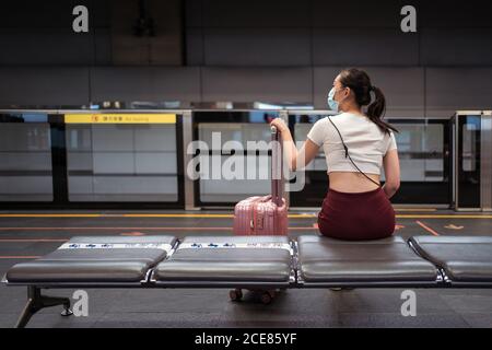 Vue arrière de la jeune femme asiatique concentrée portant une mini jupe et masque de visage assis sur un banc avec une valise en attente le train dans la gare Banque D'Images