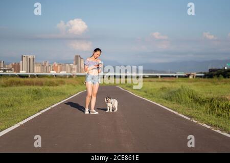 Placez la femme asiatique dans des vêtements décontractés surfer sur Internet sur un téléphone portable tout en étant debout sur une route vide avec l'américain Cocker Spaniel près de l'herbe sous le ciel nuageux en été dans la ville Banque D'Images